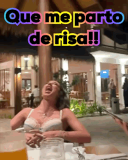 A Woman Is Sitting At A Table With A Glass Of Beer And A Sign That Says Que Me Parto De Risa !! GIF