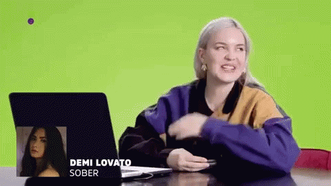 A Woman Is Sitting At A Table With A Laptop And A Picture Of Demi Lovato . GIF