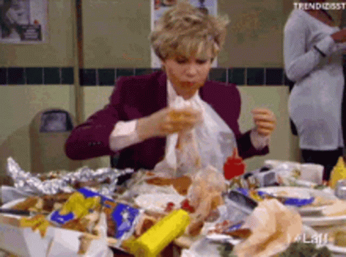 A Woman Is Sitting At A Table With A Lot Of Food On It Eating A Sandwich . GIF