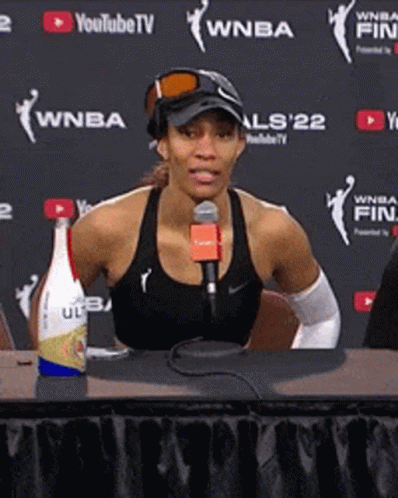 A Woman Is Sitting At A Table With A Microphone In Front Of A Wall That Says Wnba GIF