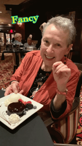 A Woman Is Sitting At A Table With A Plate Of Dessert In Front Of A Sign That Says Fancy GIF
