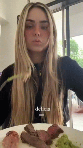 A Woman Is Sitting At A Table With A Plate Of Food And The Word Delicia Is On The Bottom Of The Picture GIF