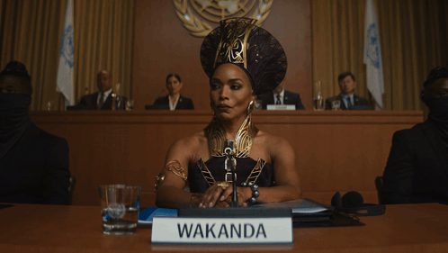 A Woman Is Sitting At A Table With A Sign That Says Wakanda GIF