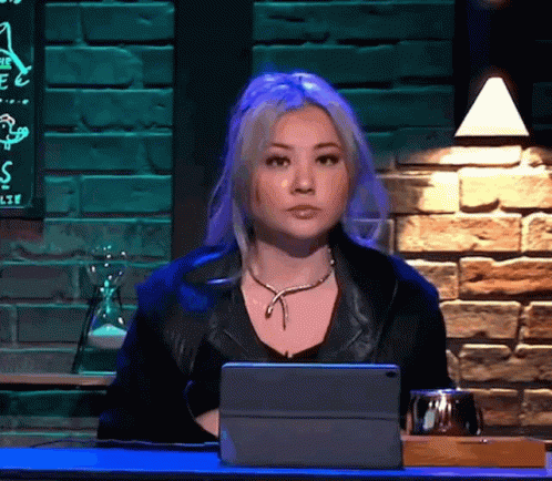 A Woman Is Sitting At A Table With A Tablet In Front Of Her And A Brick Wall Behind Her GIF