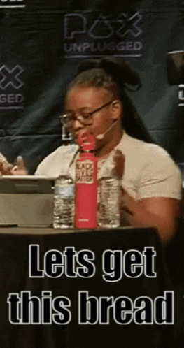 A Woman Is Sitting At A Table With A Water Bottle And A Sign That Says Lets Get This Bread GIF