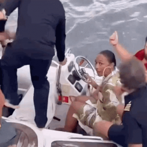 A Woman Is Sitting At The Steering Wheel Of A Boat While A Man Stands Next To Her . GIF