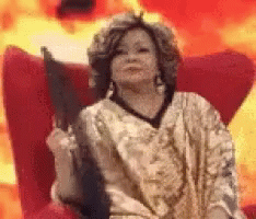 A Woman Is Sitting In A Chair Holding A Fan In Front Of Flames . GIF
