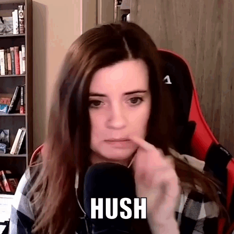 A Woman Is Sitting In A Chair In Front Of A Microphone And Making A Hush Gesture . GIF