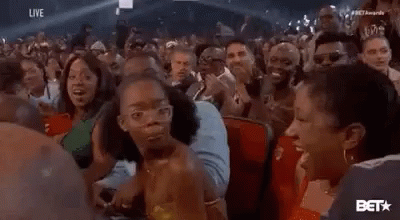 A Woman Is Sitting In A Crowd Of People At A Concert And Making A Funny Face . GIF