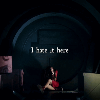 A Woman Is Sitting In A Dark Room With The Words I Hate It Here Written Above Her GIF