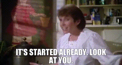 A Woman Is Sitting In A Kitchen With The Words `` It 'S Started Already Look At You '' Written On The Screen . GIF