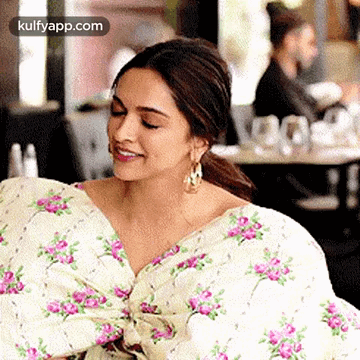 A Woman Is Sitting In A Restaurant Wearing A Floral Dress And Smiling . GIF