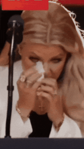 A Woman Is Sitting In Front Of A Microphone Holding A Napkin To Her Nose GIF