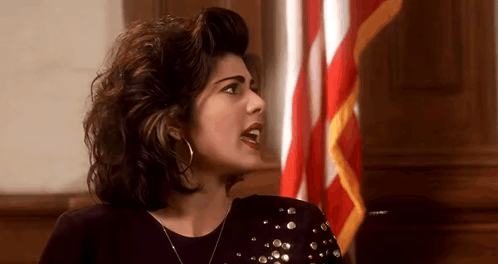 A Woman Is Sitting In Front Of An American Flag In A Courtroom . GIF