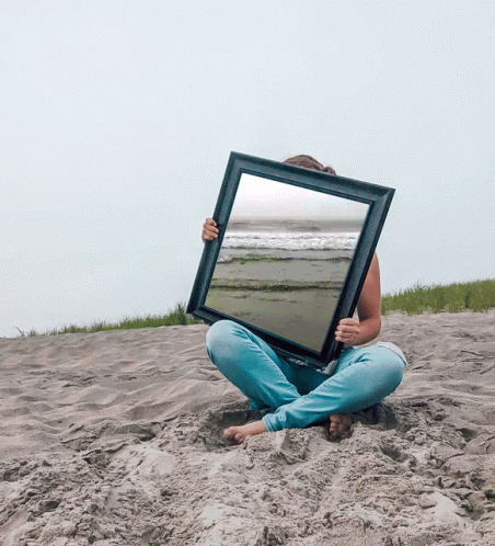 A Woman Is Sitting In The Sand Holding A Picture Frame With A Beach Scene In It GIF