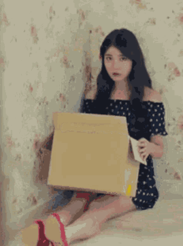 A Woman Is Sitting On A Bed Holding A Cardboard Box And A Piece Of Paper . GIF