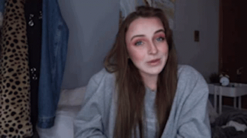 A Woman Is Sitting On A Bed Wearing A Grey Sweater And Smiling . GIF