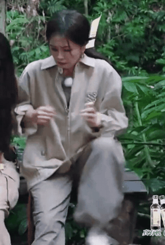 A Woman Is Sitting On A Bench In The Woods Holding A Bottle . GIF