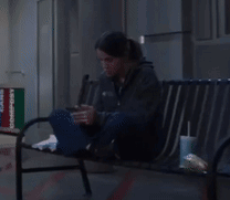 A Woman Is Sitting On A Bench Looking At Her Cell Phone GIF