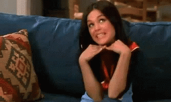 A Woman Is Sitting On A Blue Couch With Her Hands On Her Face And Smiling . GIF