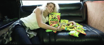 A Woman Is Sitting On A Couch Holding A Bag Of Sour Patch Kids GIF