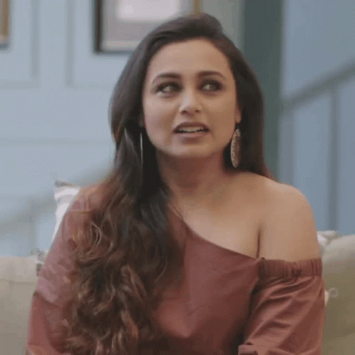 A Woman Is Sitting On A Couch Wearing A Red Off The Shoulder Top And Earrings . GIF