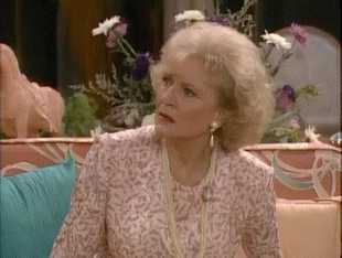 A Woman Is Sitting On A Couch With Flowers In The Background And Looking At The Camera . GIF