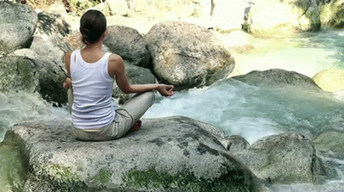 A Woman Is Sitting On A Rock In Front Of A River In A Lotus Position . GIF