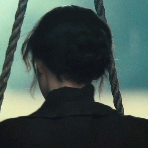 A Woman Is Sitting On A Rope Swing Looking Out Over The Ocean GIF