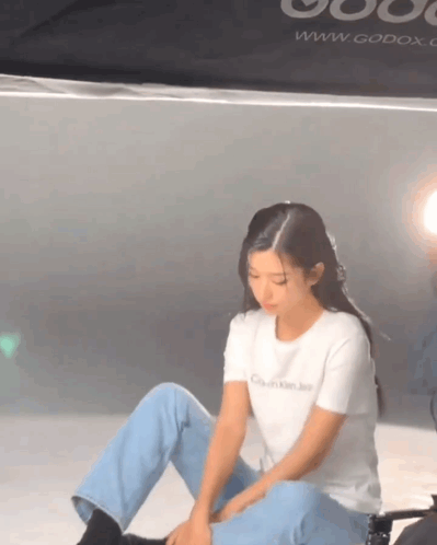 A Woman Is Sitting On The Floor Wearing A White T-Shirt That Says Calvin Klein Jeans . GIF