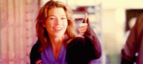 A Woman Is Smiling And Giving A Thumbs Up Sign While Standing In Front Of A Brick Wall . GIF
