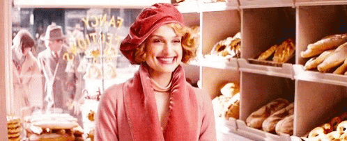 A Woman Is Smiling In A Bakery While Wearing A Red Hat And A Pink Coat . GIF