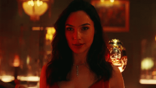 A Woman Is Smiling In A Dark Room With A Glass In The Background GIF