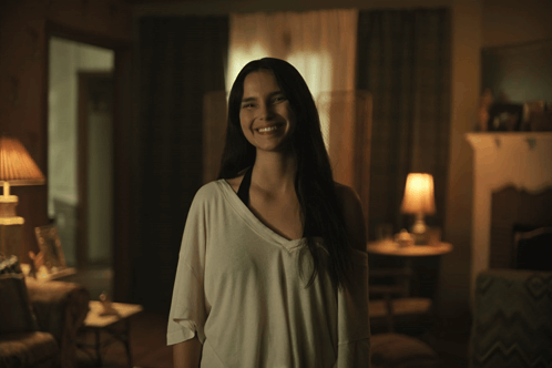 A Woman Is Smiling In A Dark Room With A Lamp In The Background GIF