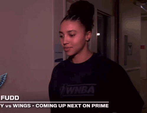 A Woman Is Smiling In Front Of A Sign That Says Fuddy Vs Wings Coming Up Next On Prime GIF