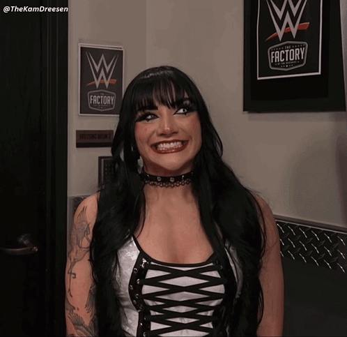 A Woman Is Smiling In Front Of A Sign That Says The Wrestling Factory GIF