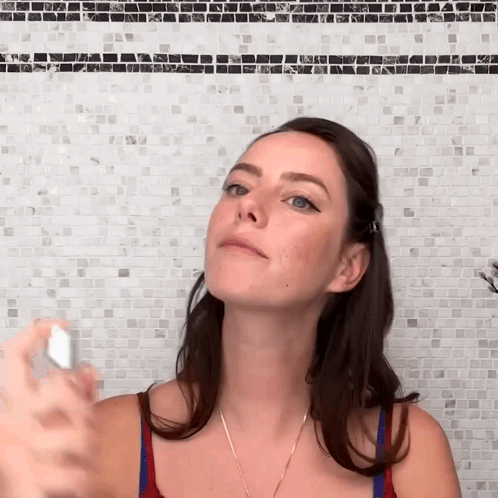 A Woman Is Spraying Something On Her Face In Front Of A Mosaic Wall GIF