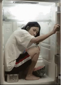 A Woman Is Squatting In An Open Refrigerator Looking For Something To Eat . GIF