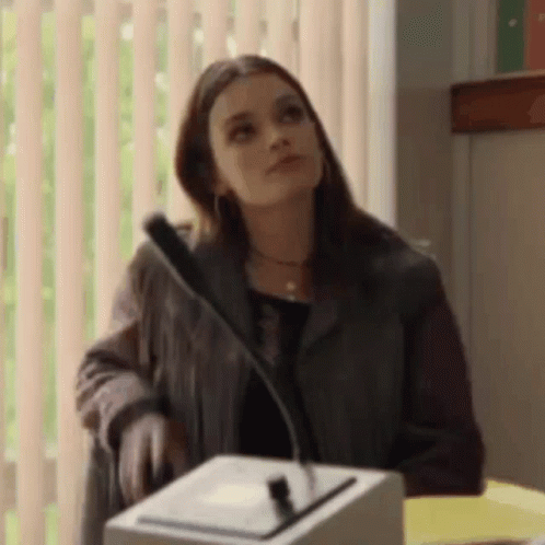 A Woman Is Standing At A Podium With A Microphone In Front Of A Window . GIF