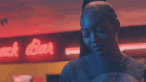 A Woman Is Standing In A Dark Room In Front Of A Neon Sign That Says Snack Bar . GIF