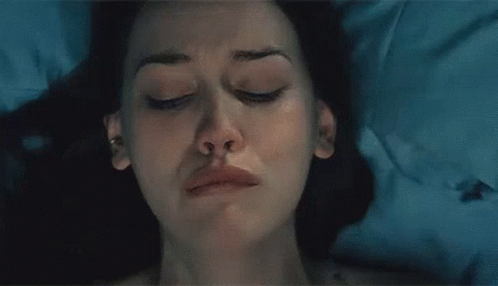 A Woman Is Standing In A Dark Room Looking Out A Window GIF