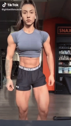 A Woman Is Standing In A Gym Wearing Under Armour Shorts And A Blue Crop Top . GIF