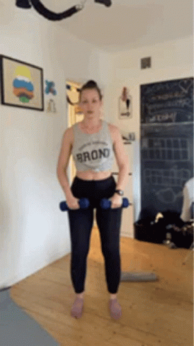 A Woman Is Standing In A Room Holding Two Dumbbells In Her Hands . GIF