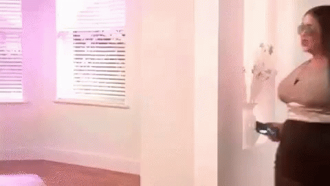 A Woman Is Standing In A Room Next To A Window Holding A Remote Control . GIF