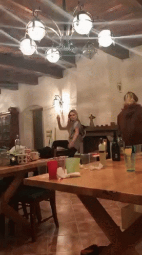 A Woman Is Standing In A Room With A Table And A Fireplace GIF