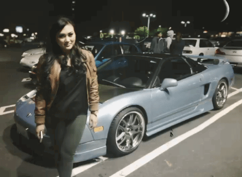 A Woman Is Standing In Front Of A Blue Sports Car With A License Plate That Says A GIF
