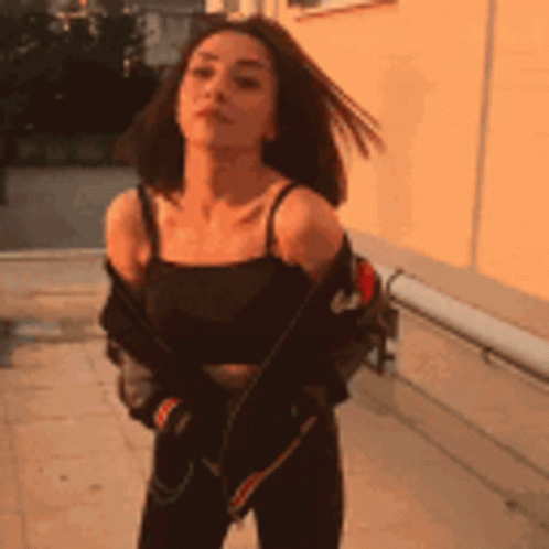 A Woman Is Standing In Front Of A Building Wearing A Black Tank Top And A Jacket . GIF