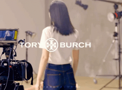 A Woman Is Standing In Front Of A Camera With Tory Burch Written On It GIF
