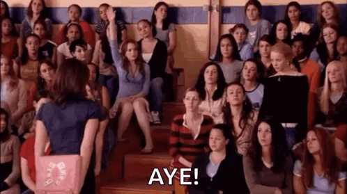 A Woman Is Standing In Front Of A Crowd Of People And Raising Her Hand To Answer A Question . GIF