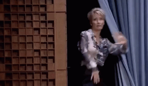 A Woman Is Standing In Front Of A Curtain And Waving Her Hand . GIF